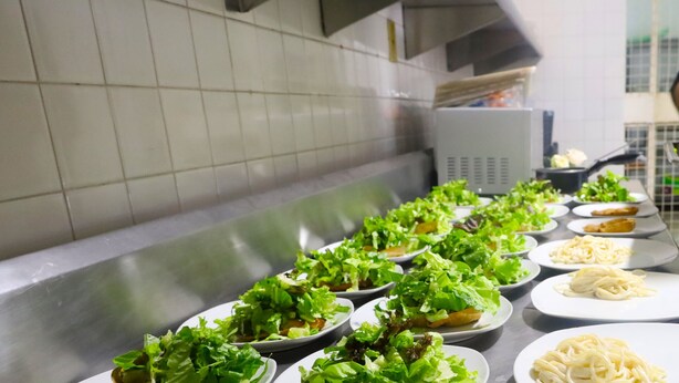 Een keuken met een roestvrijstalen aanrecht waarop borden met pasta en salades staan, en een persoon in een schort eten bereidt.