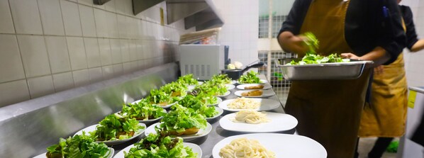 A kitchen with a stainless steel counter displaying plates of pasta and salads, and a person in an apron preparing food. - Hotel Grand Vista Cuernavaca (Cuernavaca)