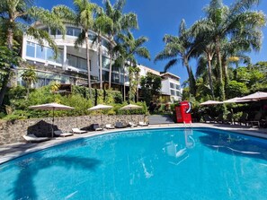 Una piscina con sillas de descanso y sombrillas, rodeada de palmeras y un edificio moderno.