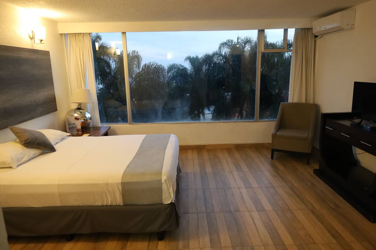 A hotel room with a bed, a desk, a chair, and a large window with a view of trees.