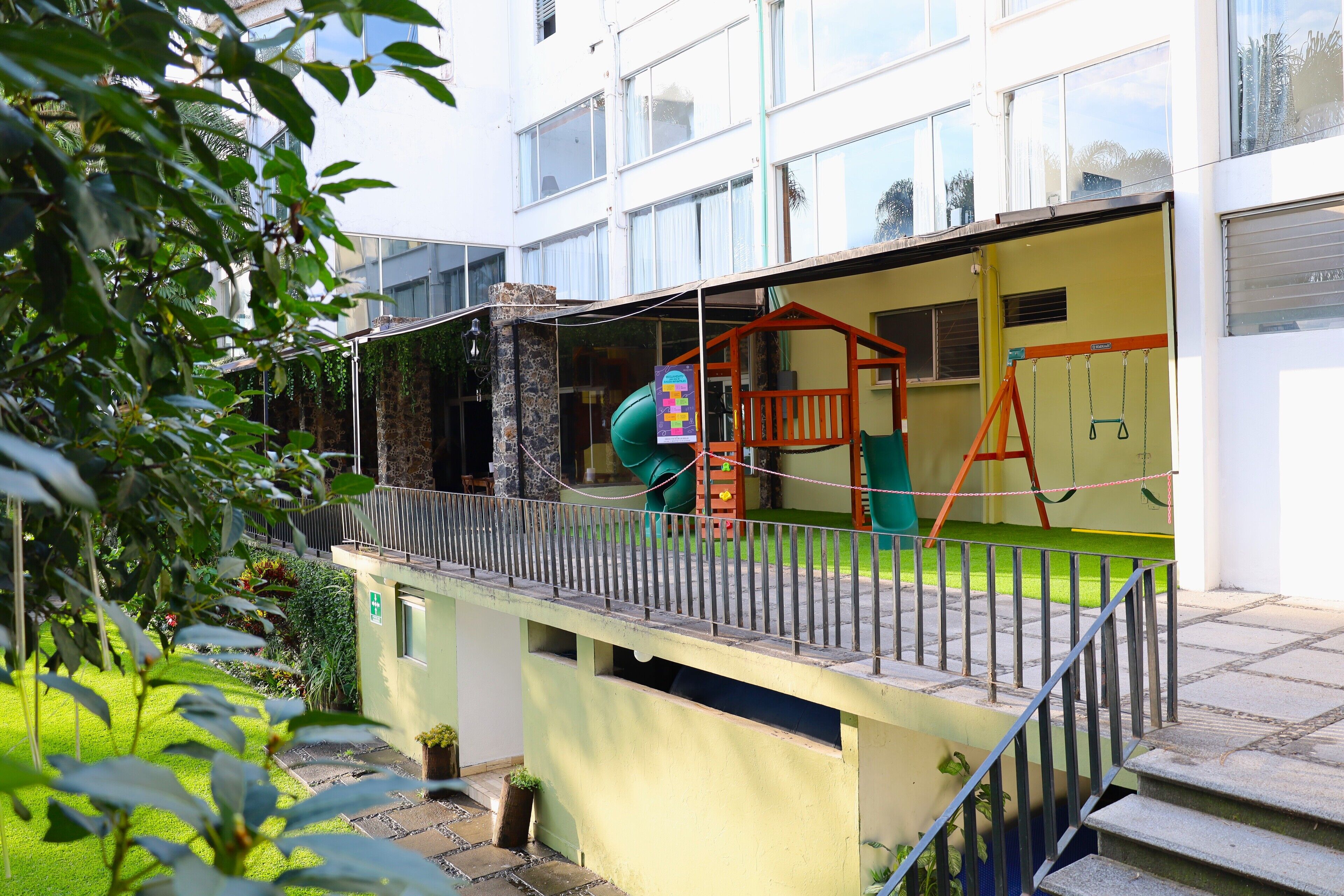Een kinderspeelplaats met een glijbaan en schommels, een kleine tuin en een gebouw met grote ramen.