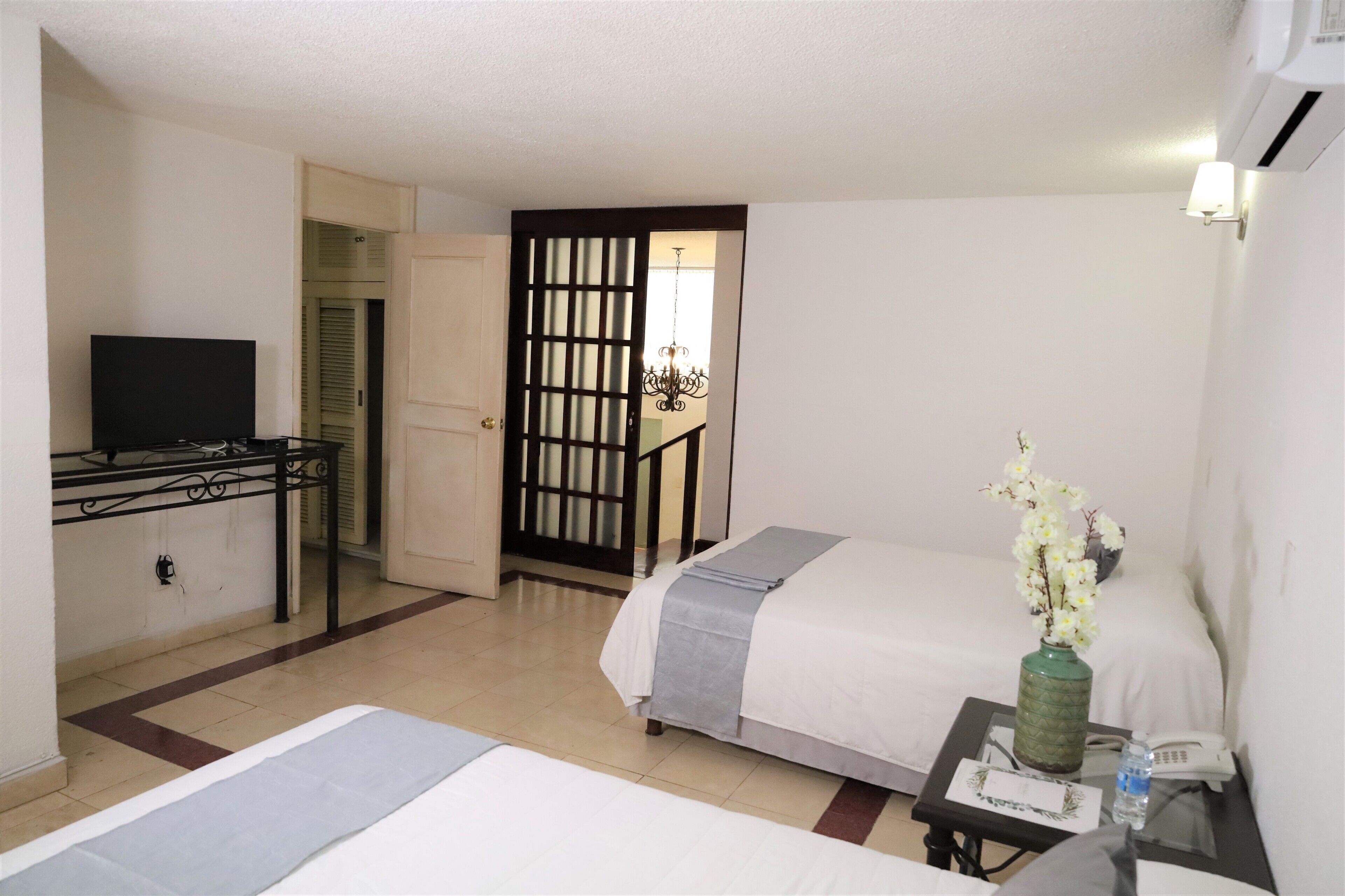 A hotel room with two beds, a TV, a desk with a phone, a vase with flowers, and a view of a staircase.