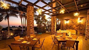 An outdoor dining area with tables and chairs, stone pillars, and a view of palm trees and a sunset. - Hotel Grand Vista Cuernavaca (Cuernavaca)