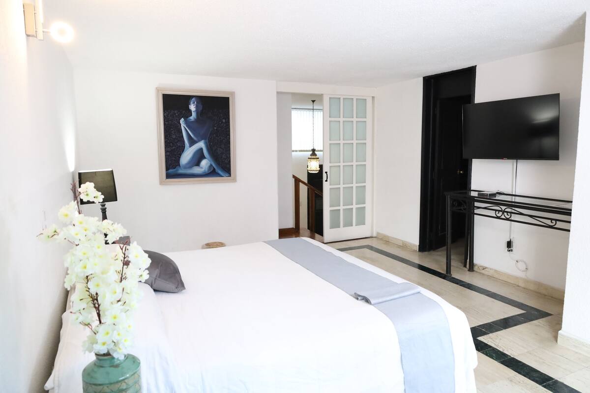 A hotel room with a bed, a TV, a painting, and a vase of flowers.