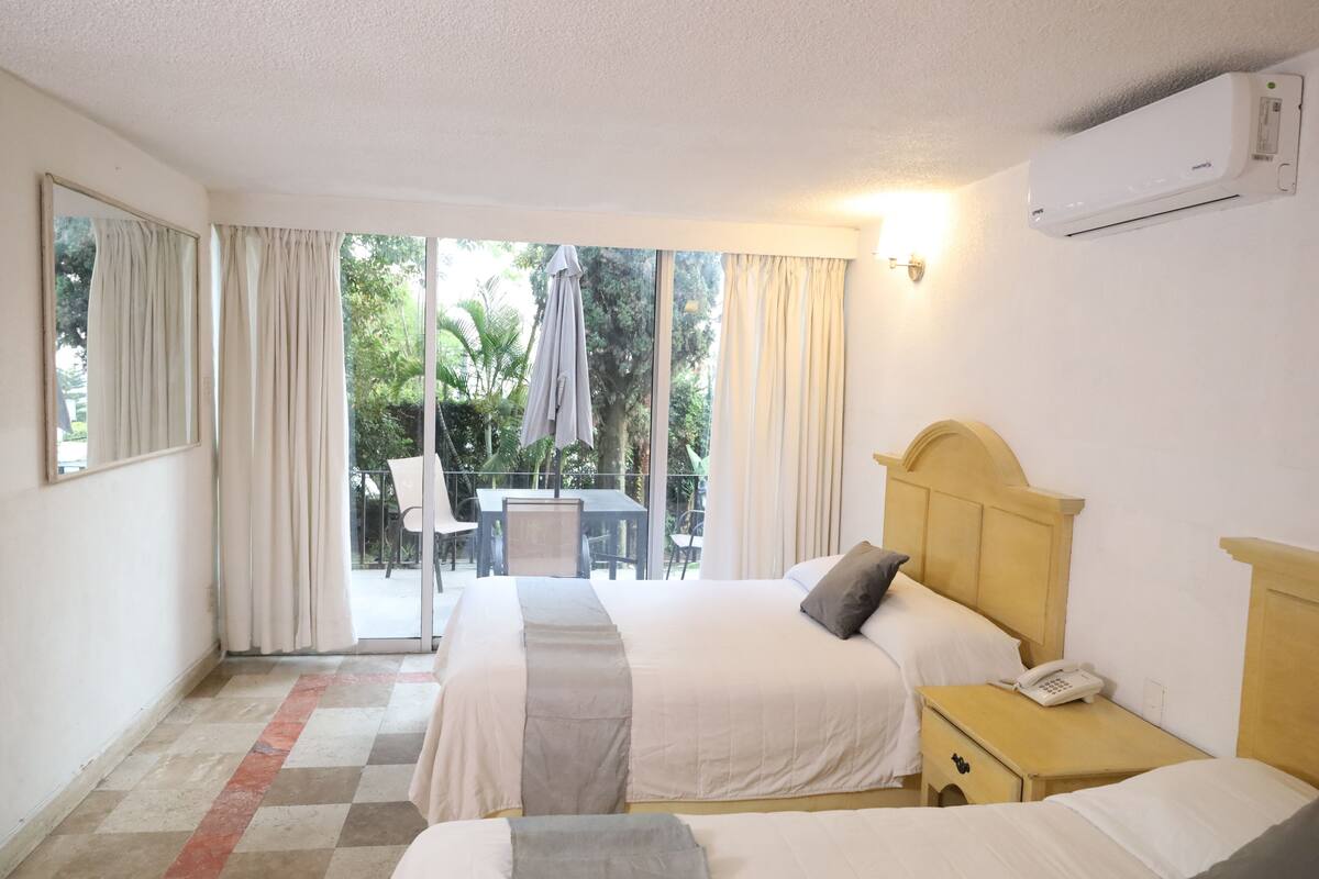 A hotel room with a large bed, a bedside table with a phone, and a view of an outdoor patio with a table and chairs.