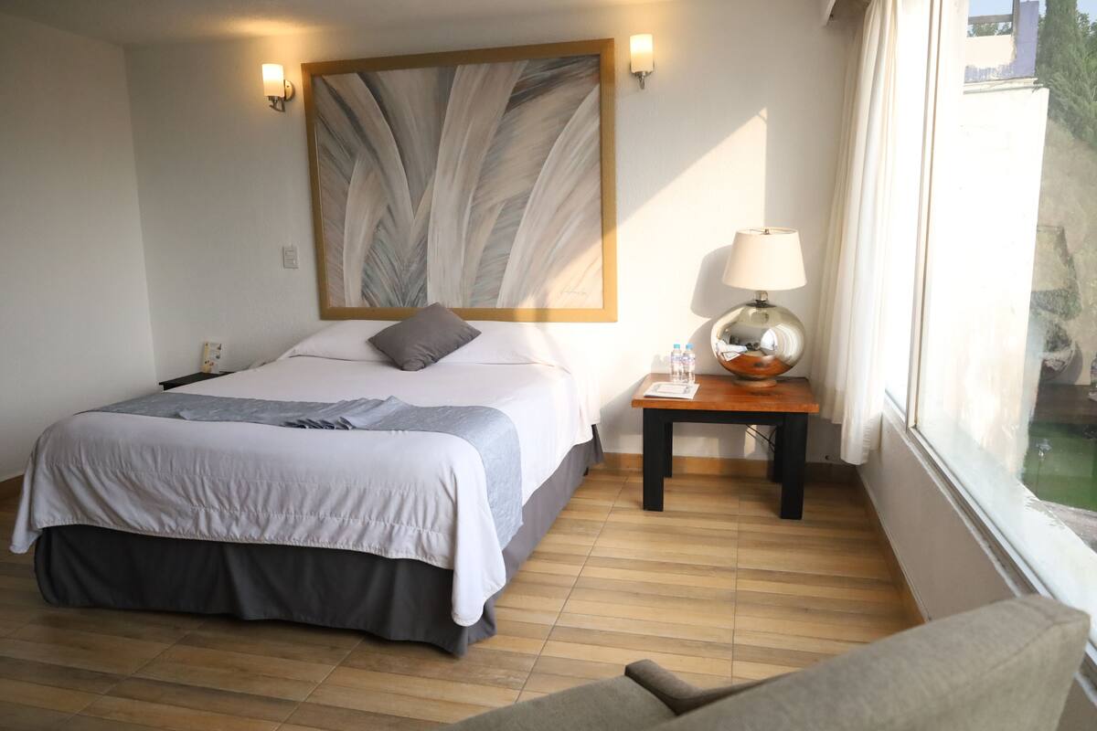A hotel room with a bed, a nightstand with a lamp, a painting on the wall, and a window with a view.