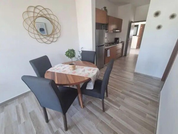 Un coin repas avec une table ronde, des chaises et une petite plante en pot. On aperçoit une cuisine à l’arrière-plan, avec des placards et un réfrigérateur.