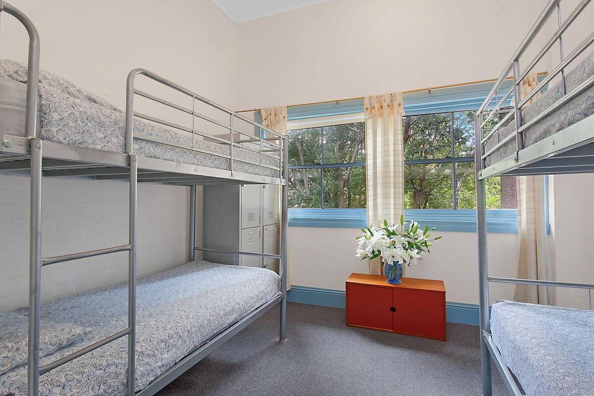 Une chambre de dortoir avec des lits superposés, une fenêtre avec des rideaux, une armoire en bois et un vase avec des fleurs.