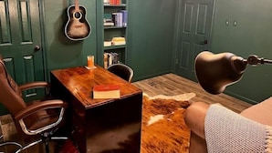 Een kamer met een houten bureau, een gitaar, een boekenplank, een vachttapijt van runderleer en een opgezette hertenkop.