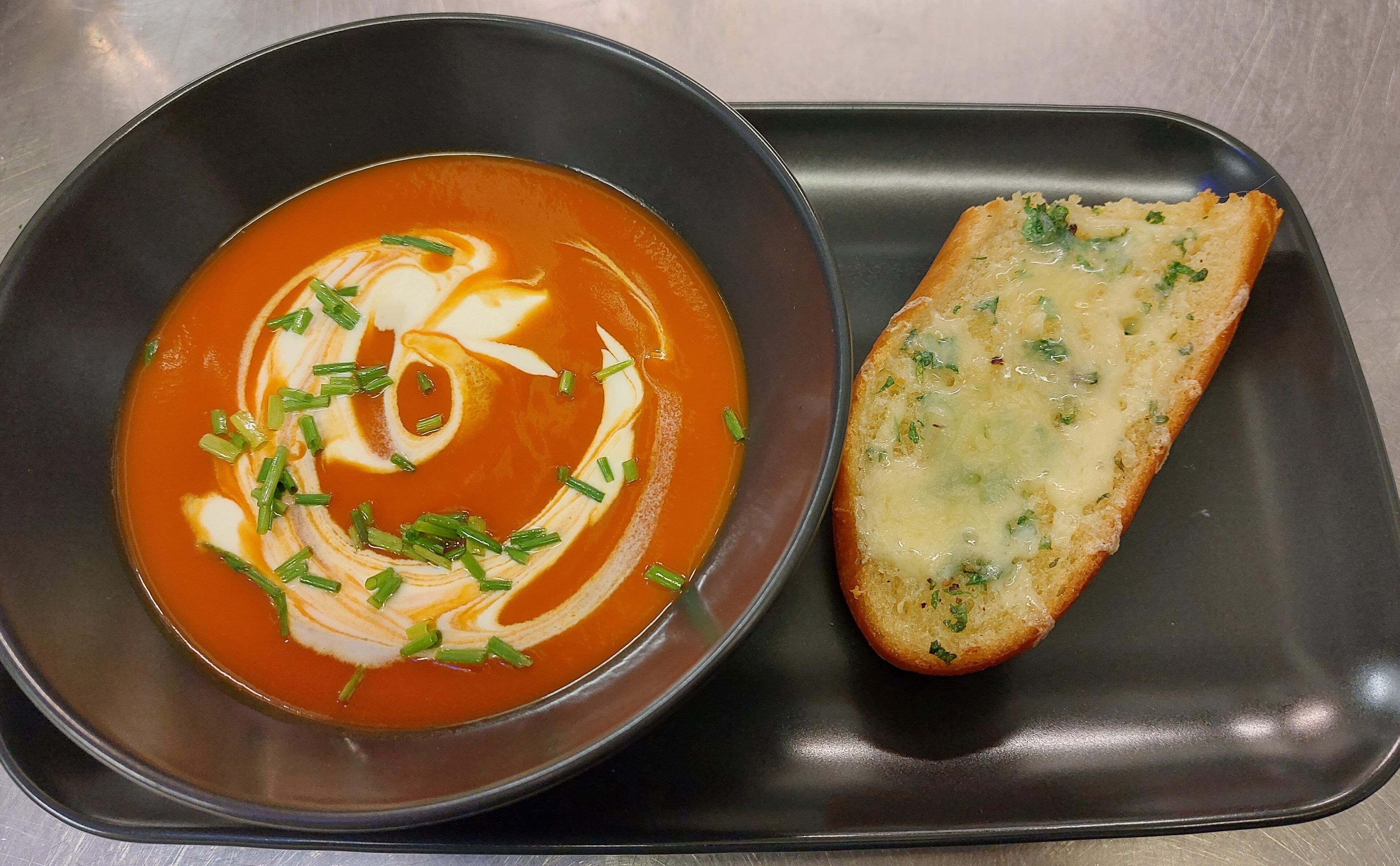 Un tazón de sopa de tomate con un remolino de crema y cebollín, acompañado de una rebanada de pan con ajo sobre una bandeja negra.