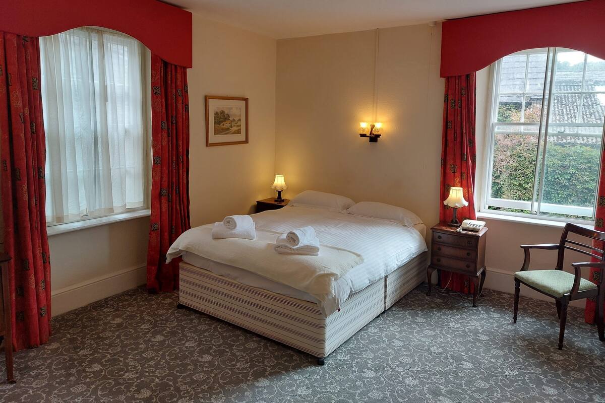 Een netjes opgemaakt bed met witte lakens en handdoeken, een bedlampje, een ingelijst schilderij aan de muur en een raam met gordijnen.
