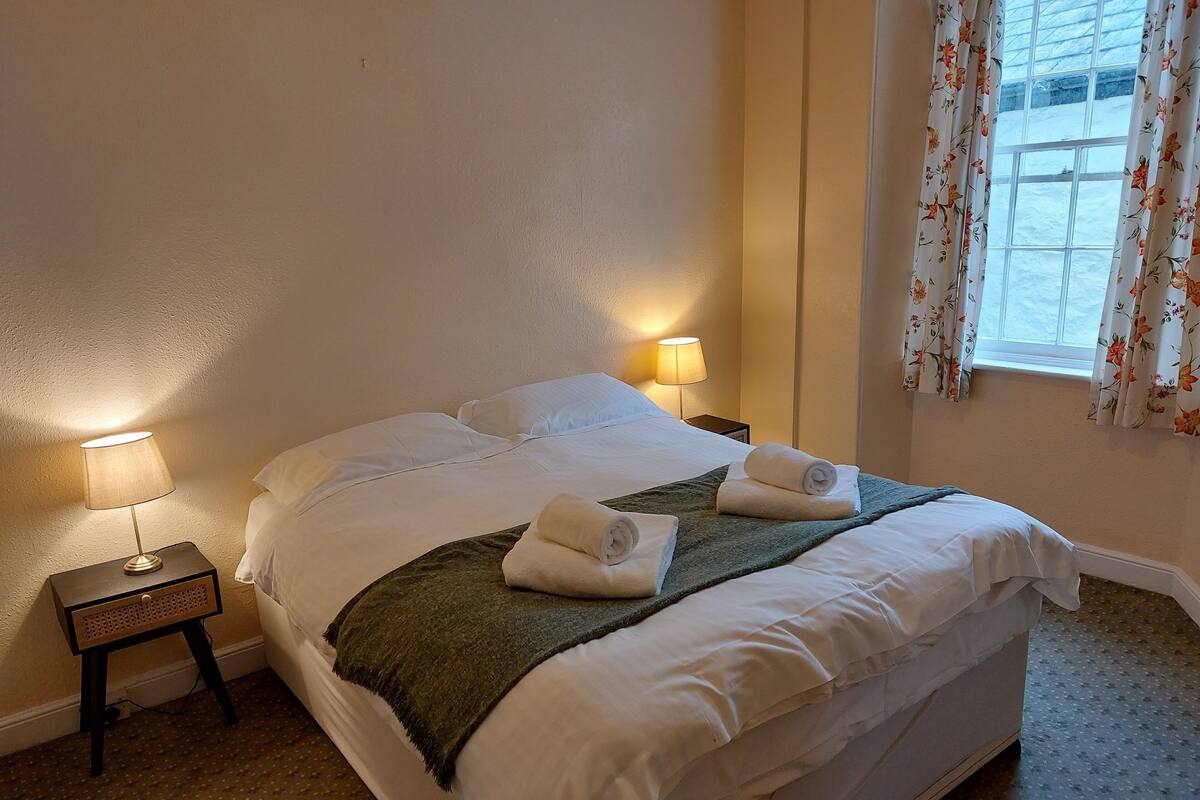 Een netjes opgemaakt bed met witte lakens en twee opgerolde handdoeken, een klein nachtkastje met een lamp en een raam met bloemen gordijnen.