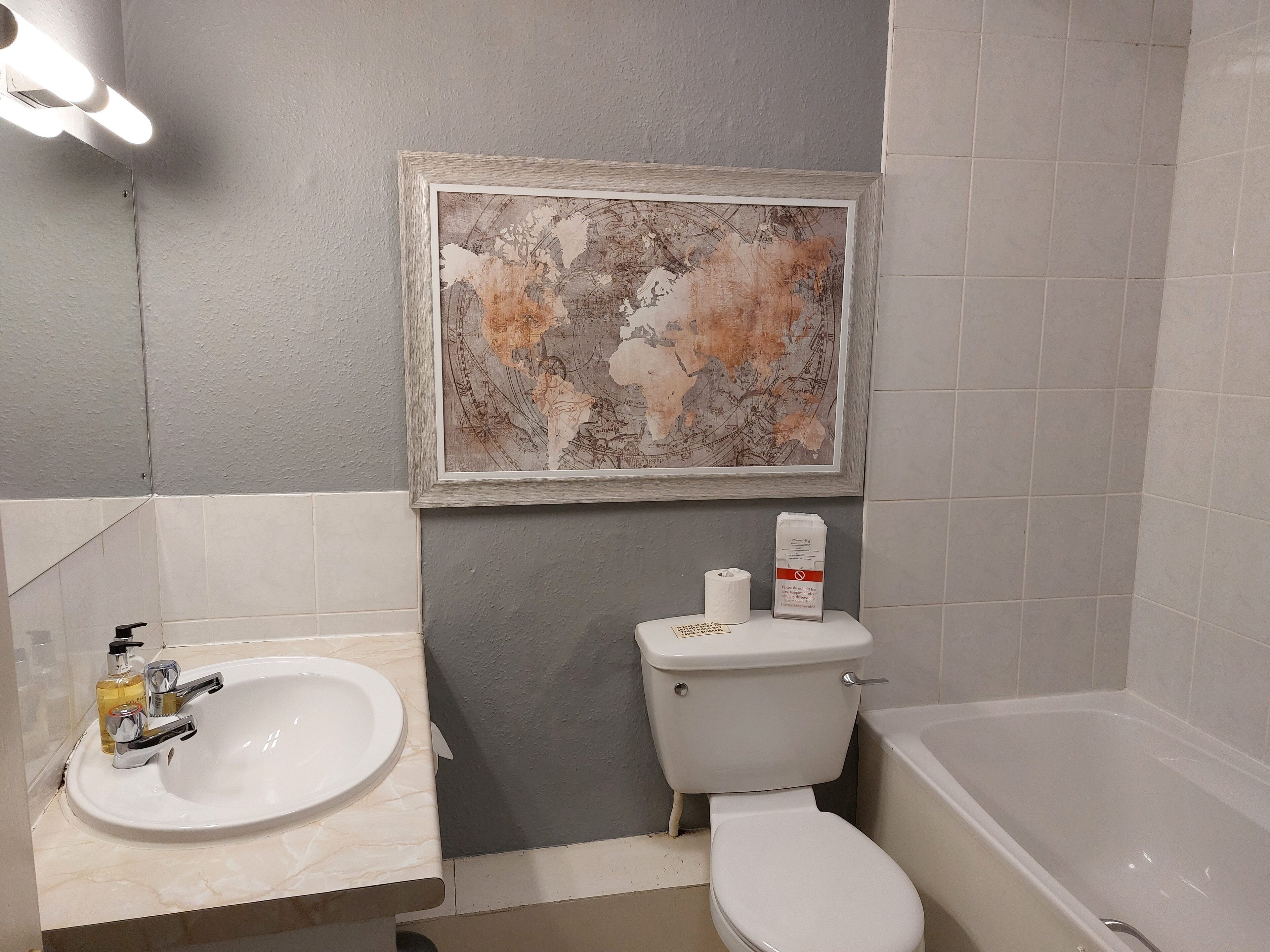 Un baño con lavabo, inodoro y bañera. Hay una obra de arte enmarcada sobre el inodoro.