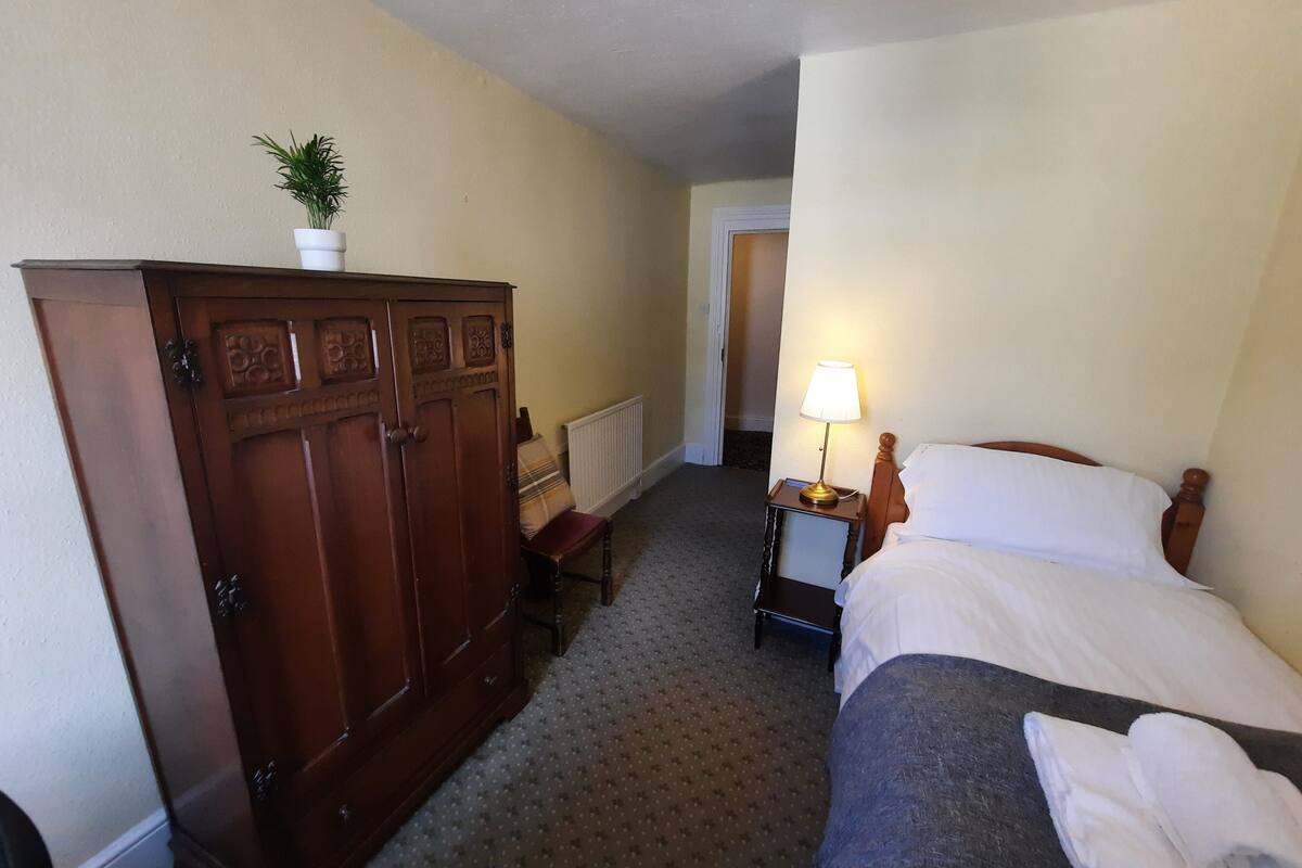 Een hotelkamer met een bed, nachtkastje met een lamp, een houten garderobe, een stoel en een potplant.