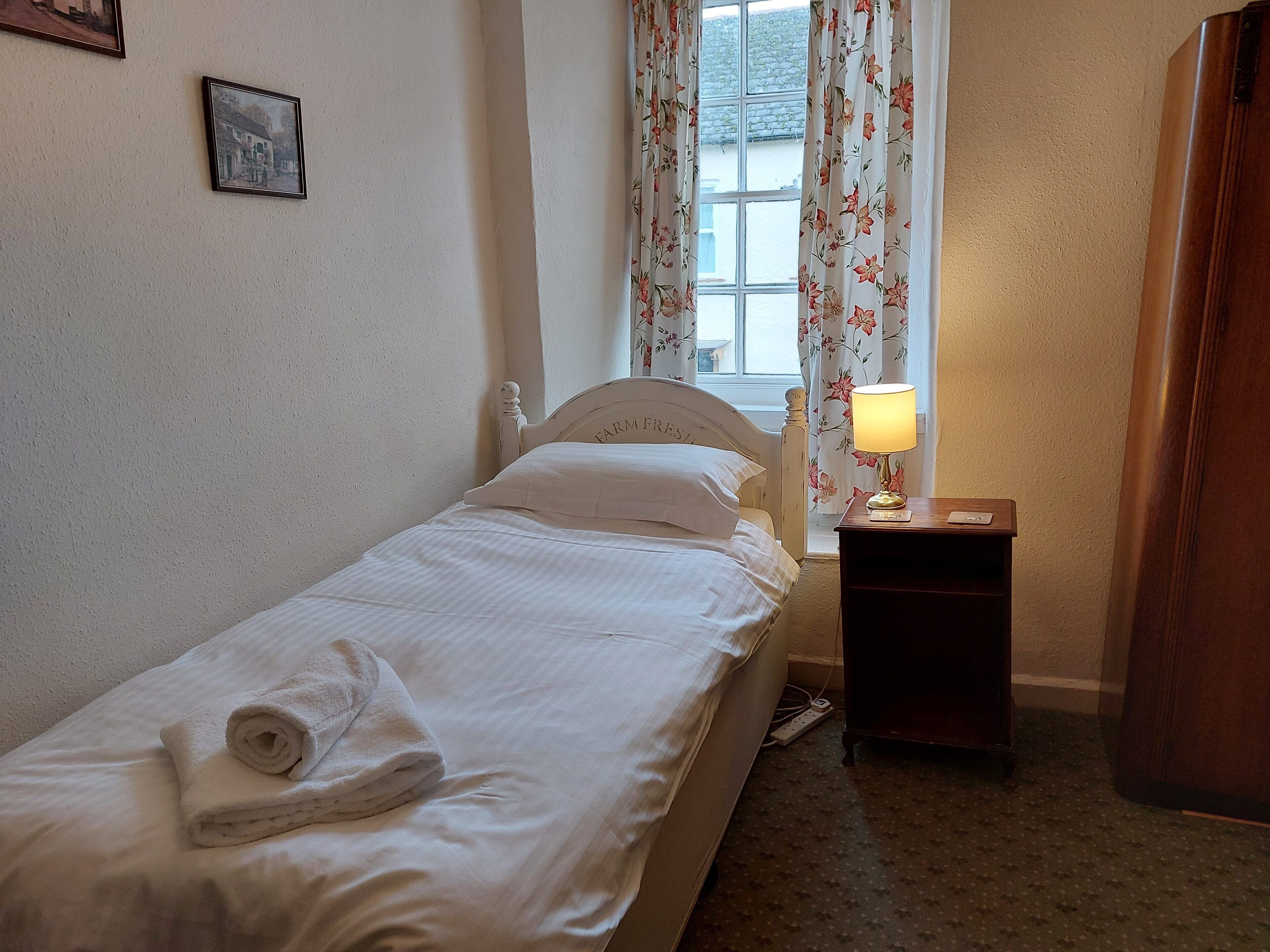 A single bed with a white headboard, a rolled towel, a nightstand with a lamp, and a window with floral curtains.