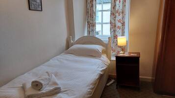 A single bed with a white headboard, a rolled towel, a nightstand with a lamp, and a window with floral curtains.