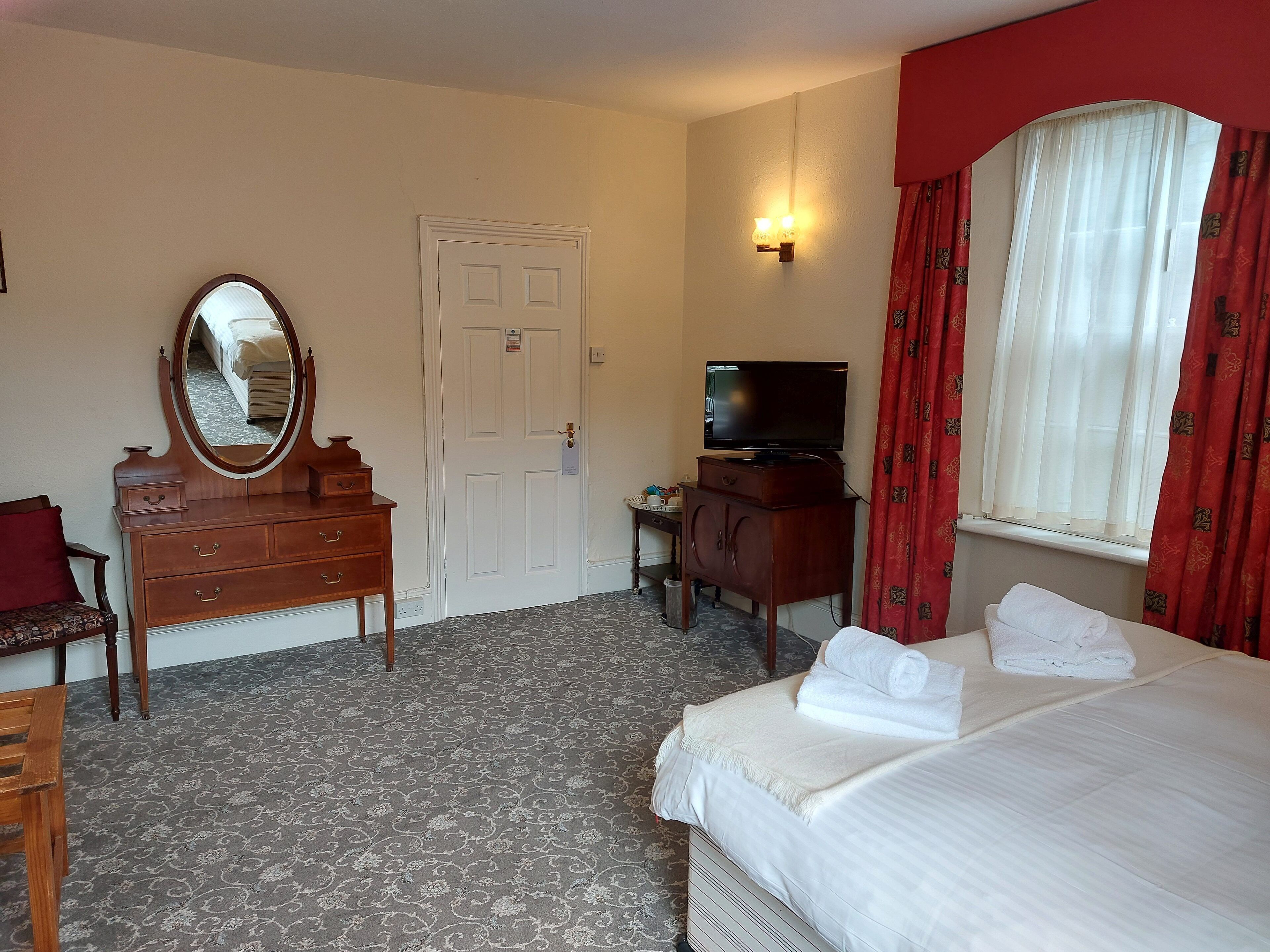A hotel room with a bed, a dresser with a mirror, a television, and a window with red curtains.