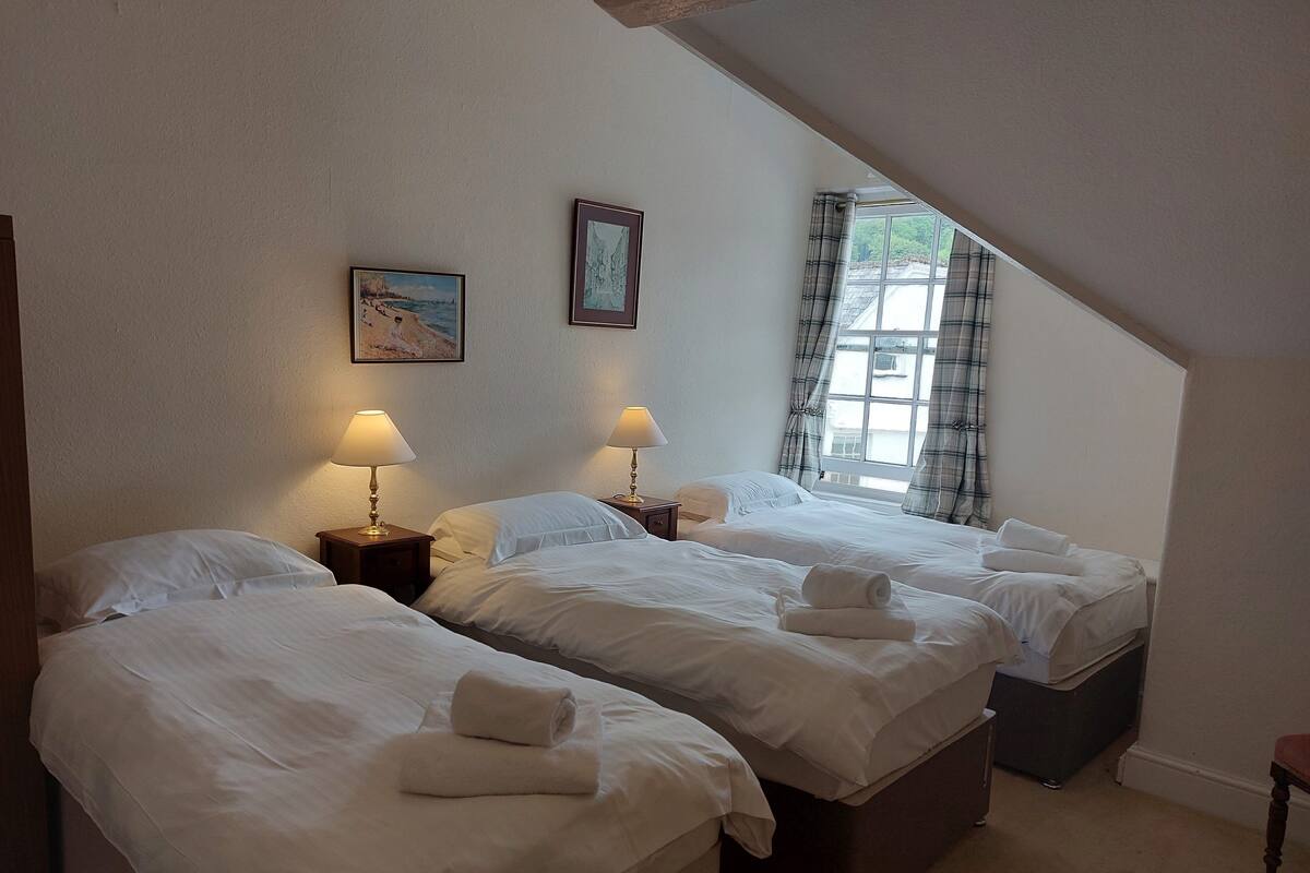 Hotelkamer met twee bedden, elk met witte lakens en een opgerolde handdoek op het bed. Twee nachtkastjes met lampen, een schilderij aan de muur en een raam met gordijnen.