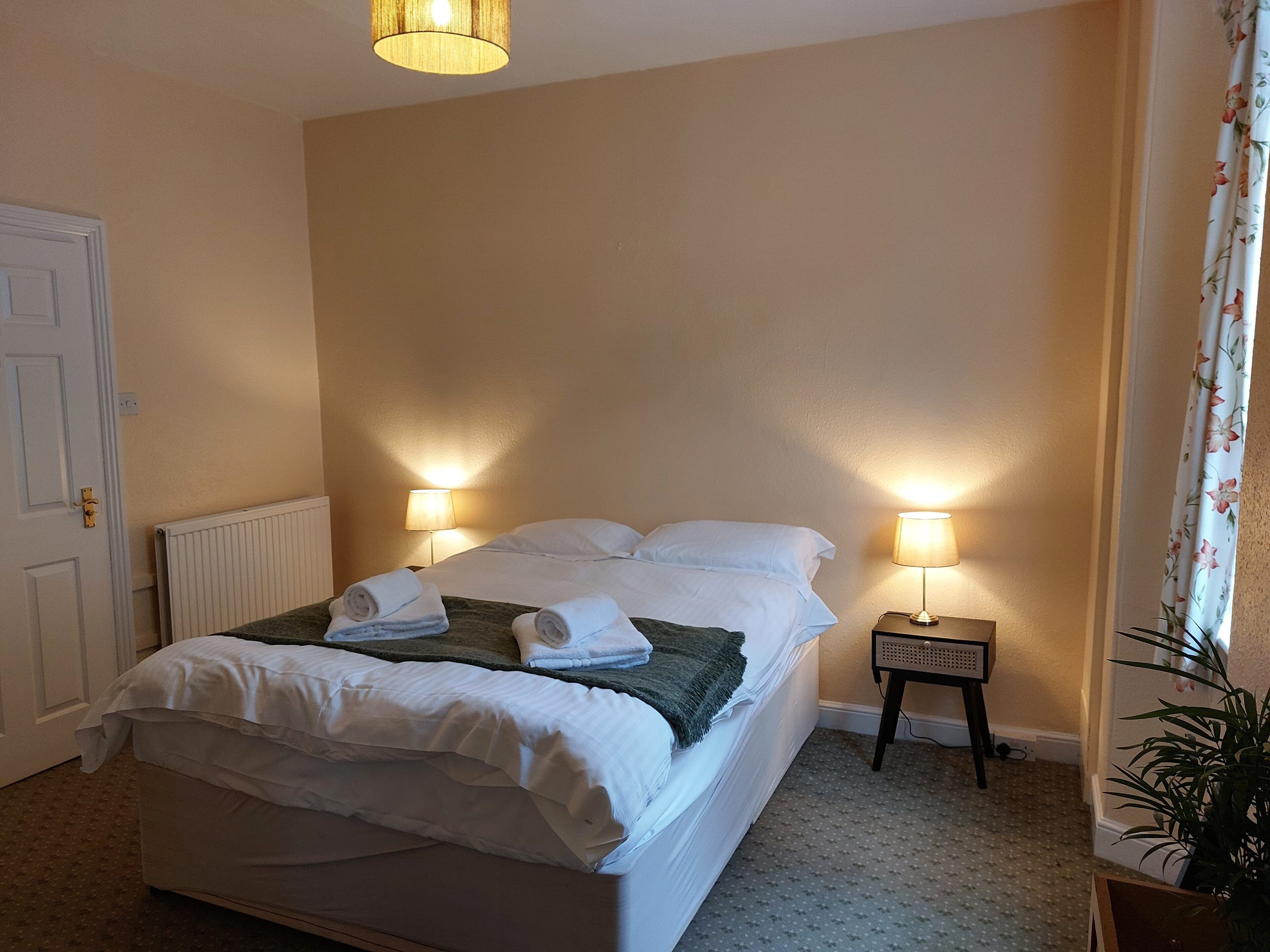 A neatly made bed with white linens and towels, two bedside lamps, a radiator, and a potted plant.