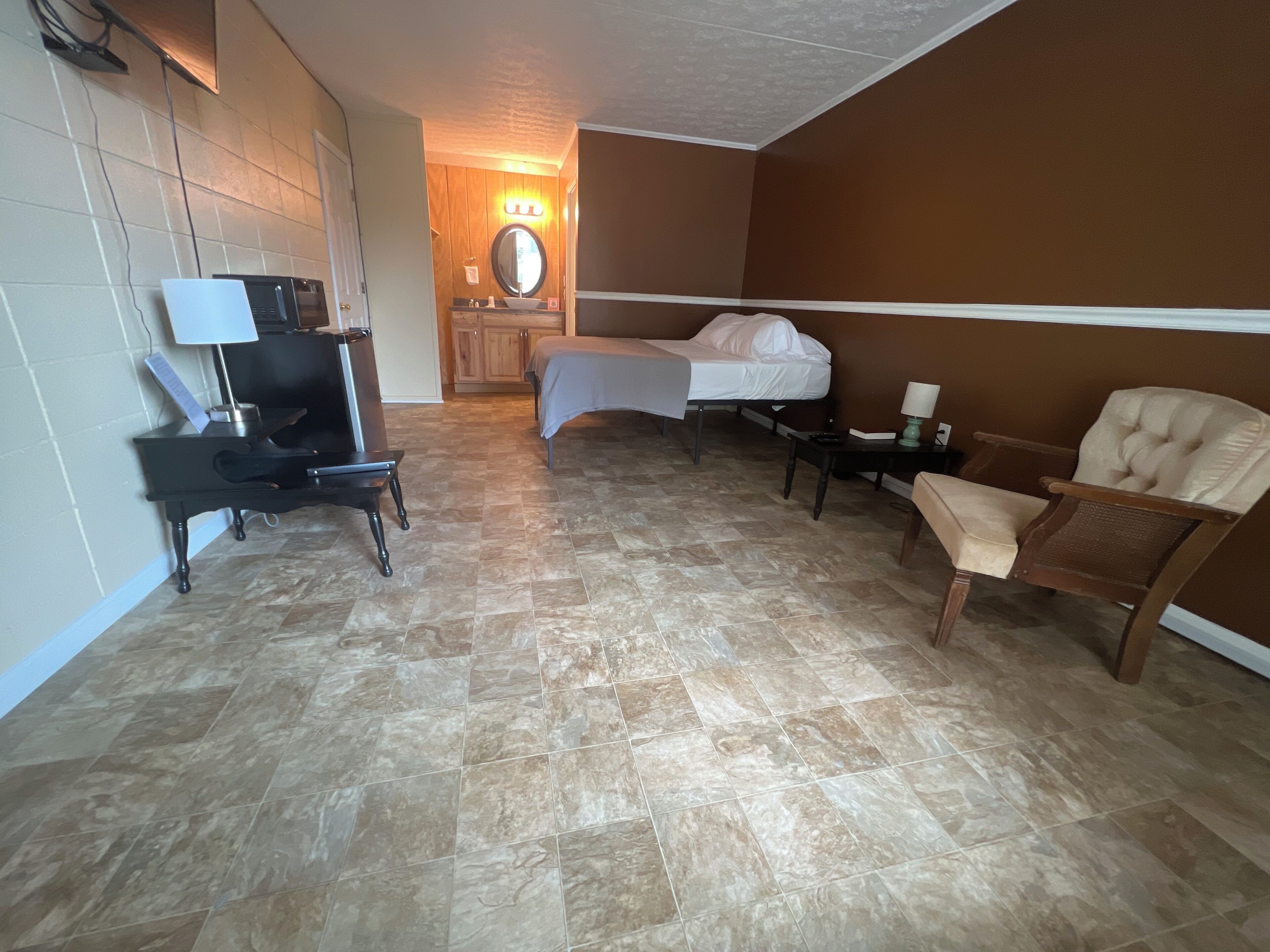 A hotel room with a bed, a chair, a desk with a lamp, a TV, and a bathroom with a mirror and sink.