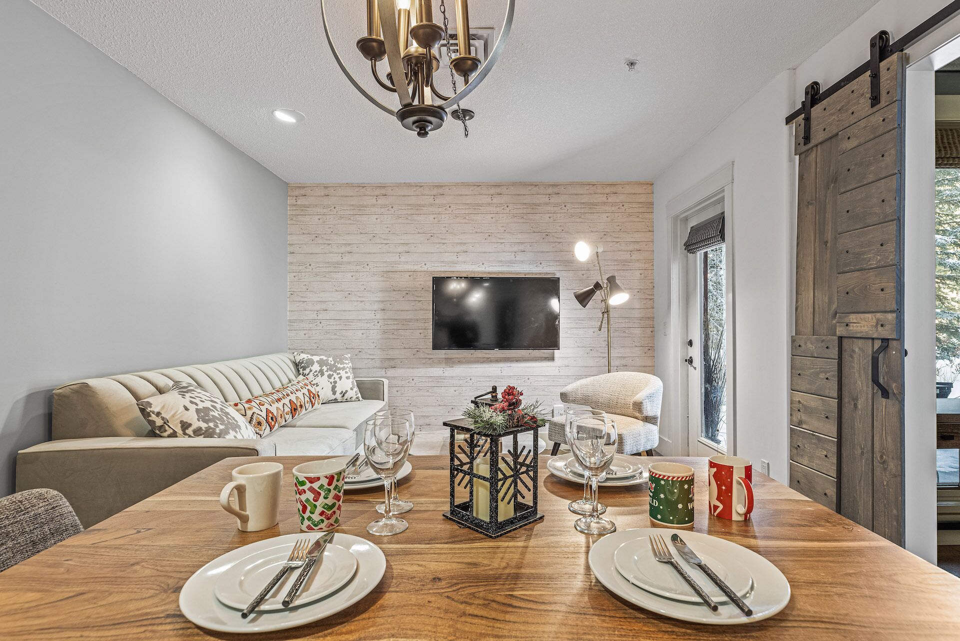 A dining area with a wooden table set for two, a sofa, a TV, and a wooden wall.