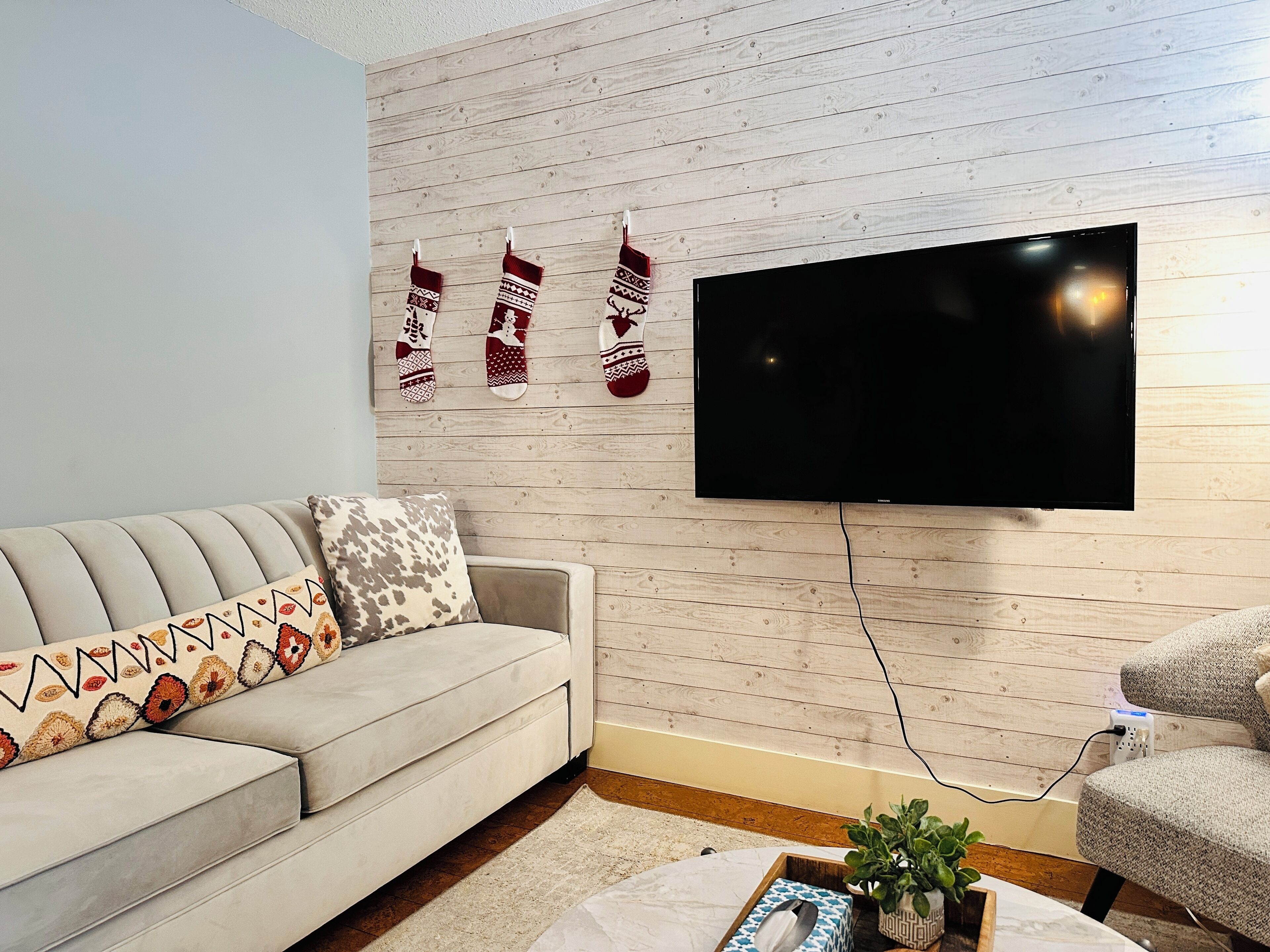 Una sala de estar con un sofá, un televisor montado en una pared de madera y un cojín decorativo estampado.