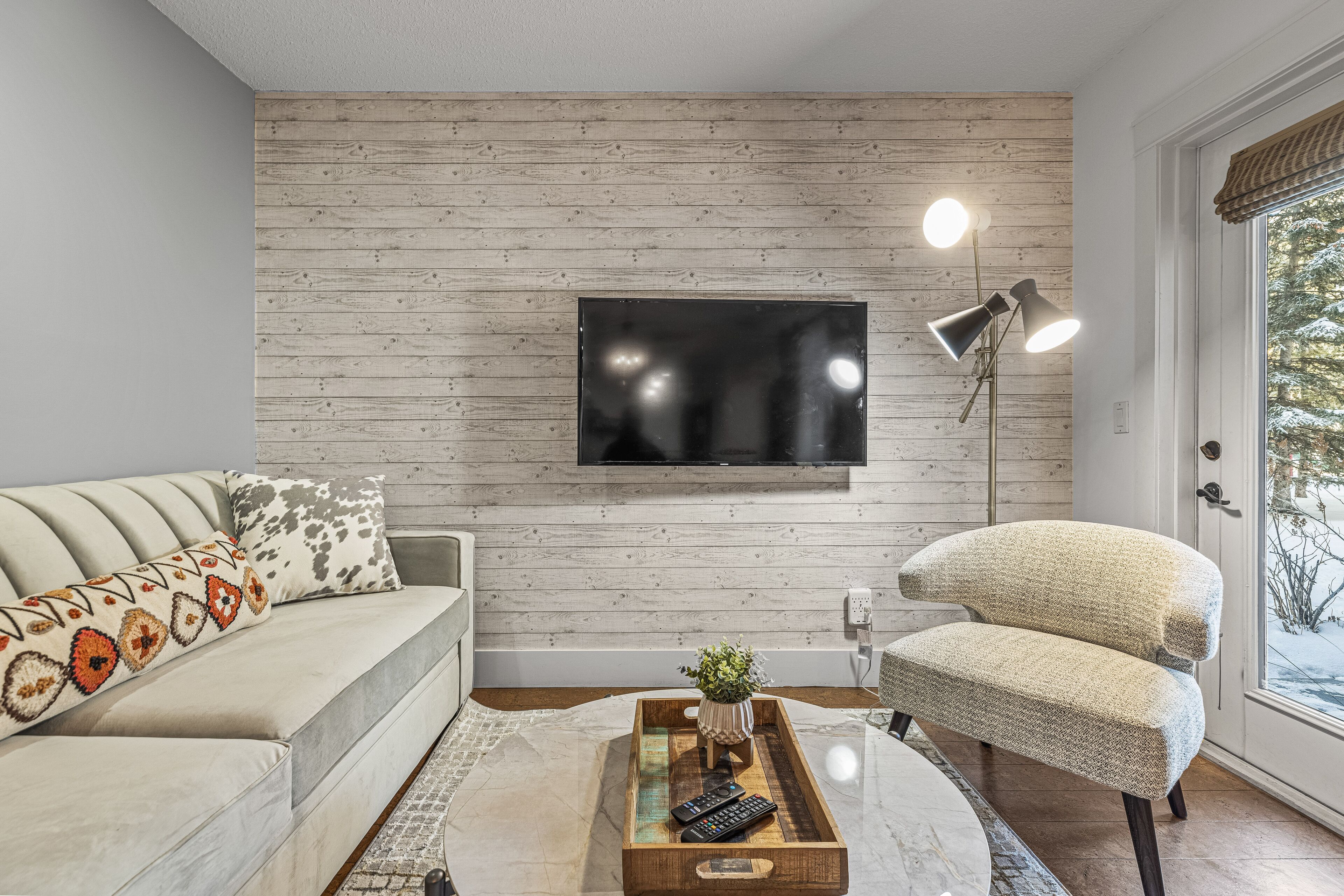 A living room with a wooden wall, a mounted TV, a sofa, a chair, a coffee table with a plant, and a floor lamp.