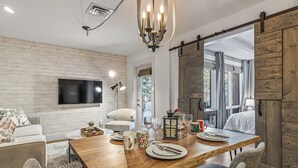 Un comedor con mesa de madera, sillas y una araña. Hay un televisor montado en la pared y una puerta corredera de estilo rústico.