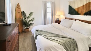 A hotel room with a bed, a wooden dresser, a surfboard, a potted plant, a lamp, and a wall art piece. - Relaxing Tropical Getaway! Coral Cottage Melbourne- family and pet friendly home (Melbourne)