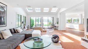 Ein großzügiger Wohnbereich mit Glastisch, grauem Sofa und weißem Pouf. Dachfenster und große Fenster sorgen für natürliches Licht.
