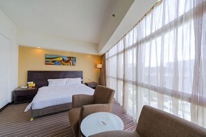 Habitación de hotel con una cama grande, dos mesitas de noche, una silla, una mesita pequeña y un ventanal con cortinas translúcidas.