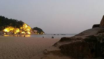 A beach with a rocky outcrop and a building with lights on the left.