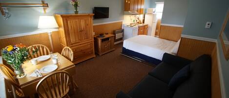 A compact hotel room with a bed, a sofa, a dining table with chairs, a kitchenette, and a ceiling fan.