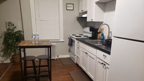 Una cocina con gabinetes blancos, una estufa y un lavamanos. Hay una mesa de madera con dos taburetes y una planta en maceta.