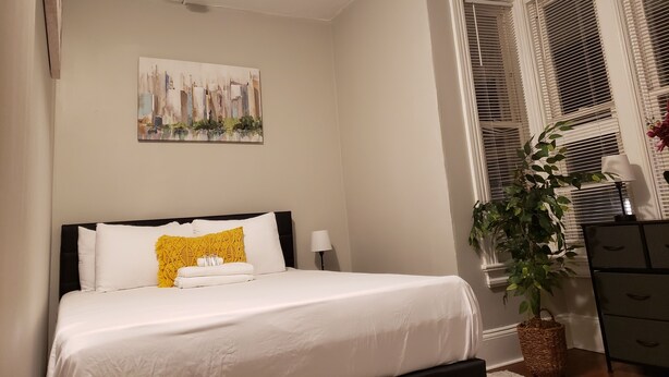 Una cama bien hecha con ropa blanca y una almohada amarilla, un cuadro de un paisaje urbano en la pared y una planta en maceta junto a la ventana.