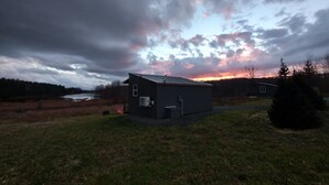 A small, single-story building with a metal roof, situated in a grassy area with a body of water in the background.