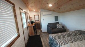 A compact room with a wooden ceiling, a bed with striped bedding, a grey sofa with a patterned pillow, a small kitchenette, and a window with blinds.