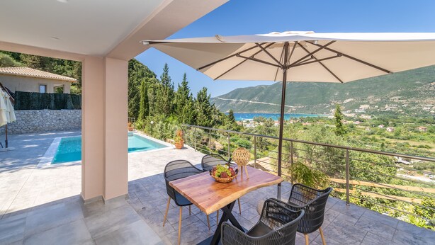 A balcony with a wooden table, two chairs, a large umbrella, and a view of a mountainous landscape. - Gyri Villa, overlooking Vassiliki bay (Lefkada)