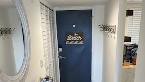 Ein Korridor mit einer dunkelblauen Tür, auf der „Beach“ steht, einem runden Spiegel, einem Holzbodenregal mit Dekorationen und einem gefliesten Boden.