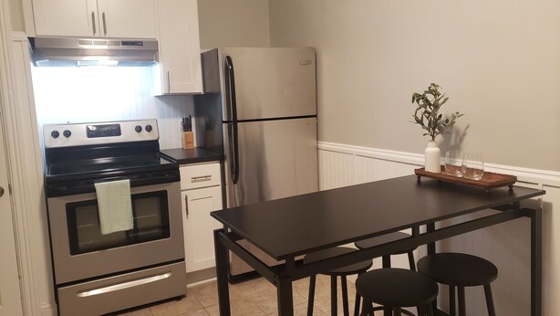 A compact kitchen with a stove, oven, refrigerator, and a small dining table with stools.