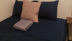A bed with navy blue bedding, a folded gray blanket, and two pillows (one navy blue, one beige). A framed picture hangs on the wall above the bed.