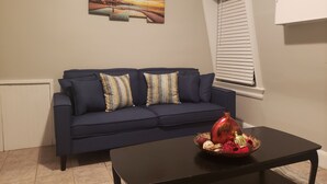 A living room with a dark blue sofa, patterned cushions, a black coffee table with decorative items, a wall-mounted painting, and a window with blinds.