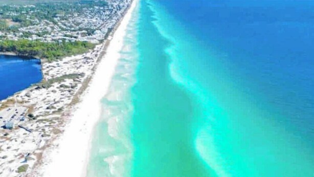 Vue aérienne d’une plage aux eaux turquoise et limpides, avec une rive sablonneuse et un petit plan d’eau sur la gauche.