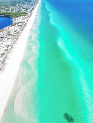 An aerial view of a beach with clear turquoise waters, a sandy shore, and a small body of water to the left.