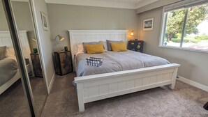 A bedroom with a white bed, a window with a view, a bedside table with a lamp, and a suitcase.