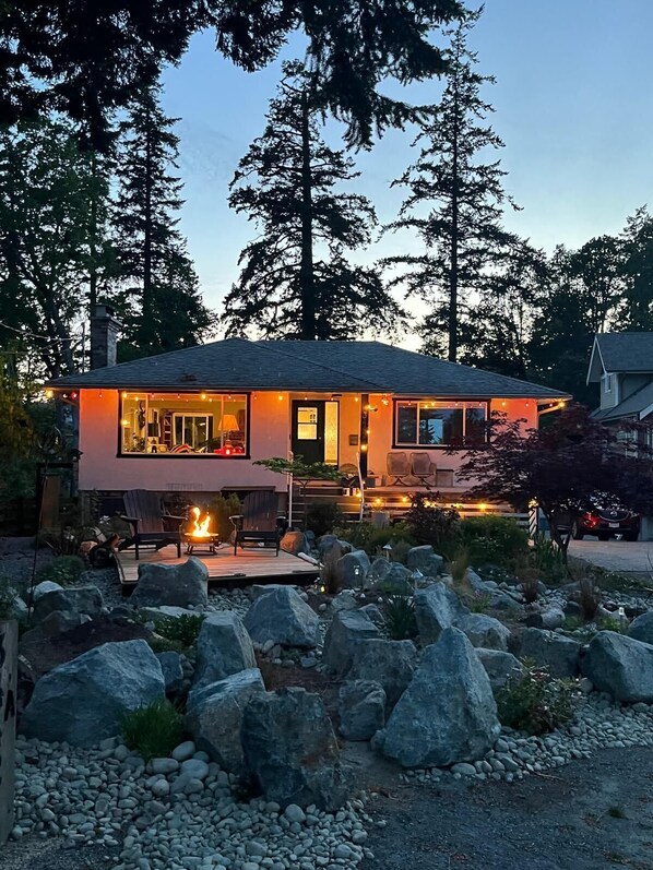 A modern house with a deck, a fire pit, and a view of tall trees.