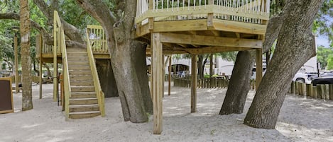 Een houten boomhuis met een trap en een platform, omgeven door bomen en zand.