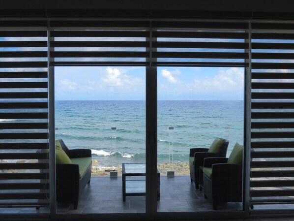A room with a view of the ocean through large windows with horizontal blinds. There are two chairs and a small table in the room.