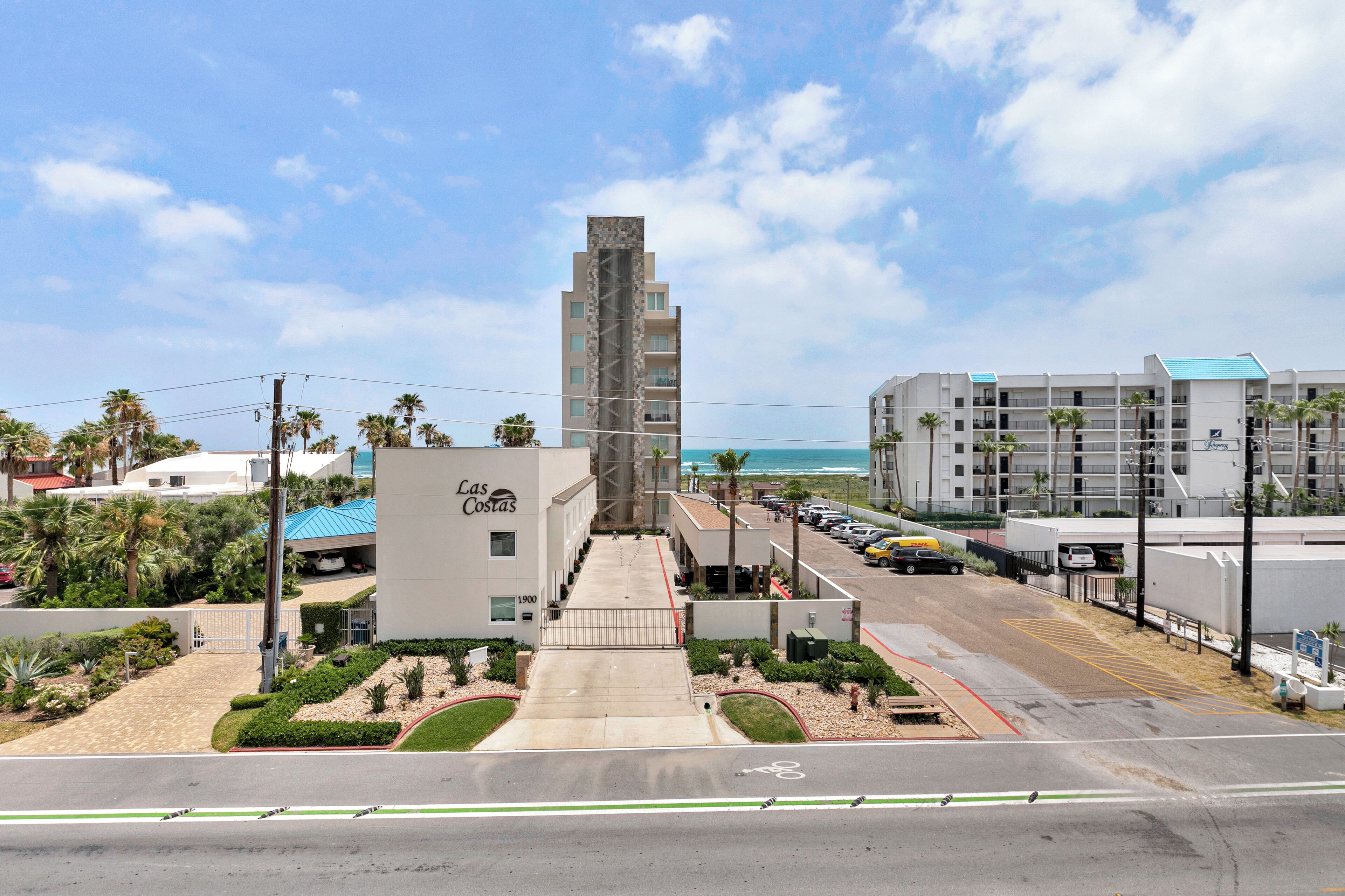 A hotel named "Las Costas" with a central tower, a parking lot, and a view of the beach.