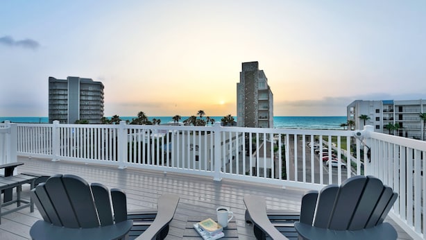 Una terraza con sillas Adirondack y una mesa, con vistas a una playa con edificios y coches aparcados.
